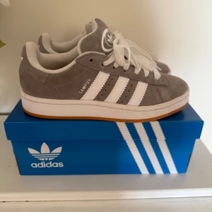 Grå Adidas Campus - Adidas Campus 00s sneakers i snygg grå mocka med klassiska vita ränder och vit sula. Skorna har rund tå, platt gummisula och vita snören. Det tillkommer även gråa snören om man vill t.ex. byta. Dem är i storlek 37 1/3, och är endast använda 2-3 gånger💕💕 Skriv för fler bilder. Pris kan diskuteras🥰