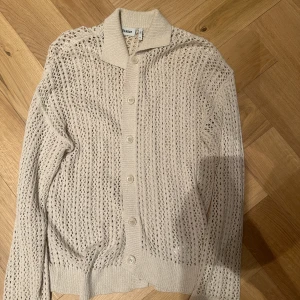 Beige stickad kofta från Weekday - Säljer en beige stickad kofta från Weekday med snyggt hålmönster och ribbade muddar. Koftan har långa ärmar, knappar framtill och en klassisk krage. Perfekt för lager-på-lager och chill stil.