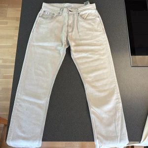 Ljusgrå jeans från Calvin Klein - Snygga ljusgrå jeans från Calvin Klein Jeans med raka ben och klassisk femficksdesign. Byxorna har silverfärgade detaljer och logoknapp framtill. Perfekta för dig som gillar stilrena och tidlösa jeans i bomull.