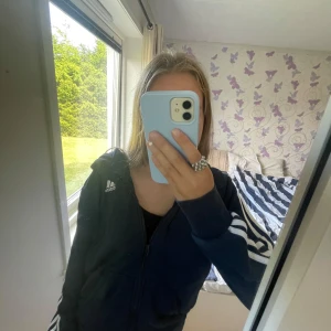 Mörkblå Adidas zip-hoodie med ränder - Säljer en mörkblå Adidas hoodie med dragkedja och huva. Klassiska vita ränder längs ärmarna och Adidas-logga på bröstet. Skön passform och perfekt för chill dagar. Materialet är mjukt och känns sportigt. 