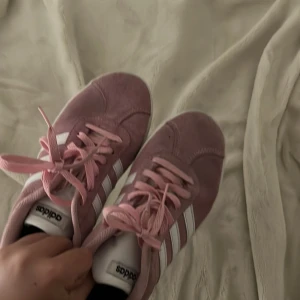 Adidas Gazelle rosa sneakers strl 35 - Säljer ett par rosa Adidas Gazelle sneakers med klassiska vita ränder och snörning. Ovandelen är i mocka och de har en platt sula. Perfekta för dig som vill ha en sportig och snygg look med en touch av färg.