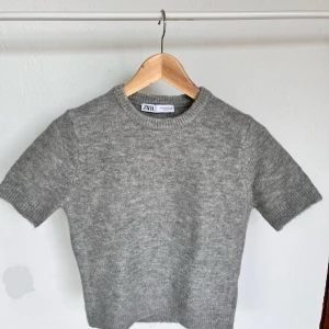 Grå stickad kortärmad tröja från Zara - Mysig grå stickad tröja från Zara med korta ärmar och rund hals. Tröjan har en enkel och clean look med ribbade muddar vid ärmslut och nederkant. Perfekt för dig som gillar basic och vill ha något som funkar till många olika stilar.