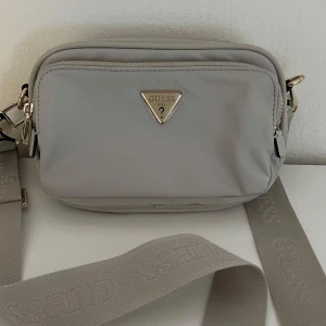 Beige axelväska från Guess - Snygg beige axelväska från Guess med justerbar bred rem och guldfärgade detaljer. Väskan har två stora fack med dragkedja och ett mindre innerfack. Guess-logga i metall framtill och diskret logotyp på remmen. Perfekt storlek för mobilen och det viktigaste.OBS väskan är original och använd 1 gång.