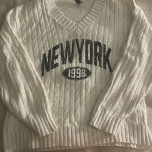 Vit stickad tröja med New York-tryck - Säljer en vit stickad tröja från H&M Divided med grov kabelstickning och svart New York-tryck framtill. Tröjan har v-ringad hals, ribbade muddar och lång ärm. Perfekt för dig som gillar sportig och urban stil.