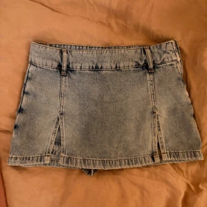 Ljusblå jeanskjol med slitsar - Säljer en ljusblå jeanskjol med två slitsar framtill och klassiska fickor bak. Kjolen har hällor för bälte och en rak, kort passform som ger en cool och avslappnad vibe. Perfekt att styla med en enkel t-shirt eller hoodie.