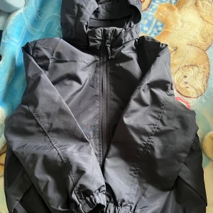 Svart vindjacka med huva från H&M - Svart vindjacka från H&M i storlek 110/116. Jackan har huva, dragkedja framtill och resår vid ärmsluten. Perfekt för blåsiga dagar och enkel att matcha med allt. Snygg och stilren design som funkar till det mesta. Aldrig använd och inga defekter. Kommer från ett djur och rökfritt hem. 