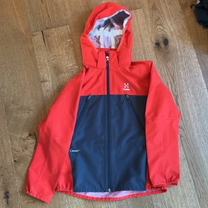 Haglöfs skaljacka 152 - Snygg tvåfärgad skaljacka från Haglöfs i rött och grått. Perfekt för regniga dagar och outdoor-äventyr. Juniorstorlek 152.