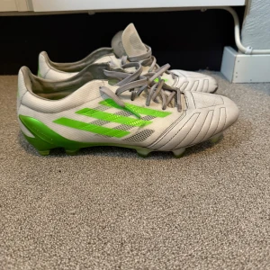 Vita och gröna Adidas fotbollsskor - Snygga fotbollsskor från Adidas i vitt med gröna detaljer och klassiska ränder på sidorna. Skorna har snörning och är tillverkade i syntetmaterial med mönstrad ovandel och dobbar för bra grepp på planen. Perfekta för dig som vill sticka ut på matchen!