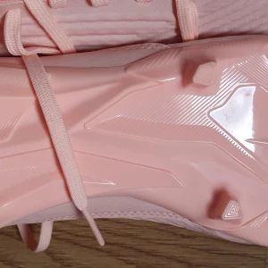 Adidas rosa fotbollsskor med snörning - Snygga fotbollsskor från Adidas i en ljusrosa färg med klassiska tre ränder på sidan. Skorna har snörning och en ovandel i syntetmaterial med textilinslag. Yttersulan har dobbar för bra grepp på planen. Perfekta för dig som vill sticka ut på matchen!. Original pris 1399 kr