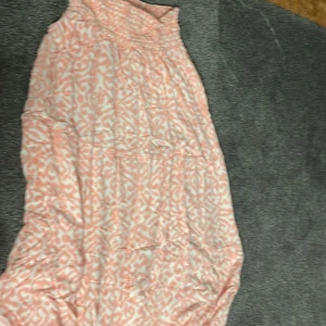 Rosa och vit kortklänning med mönster - Säljer en söt kortklänning i rosa och vitt med ett unikt mönster. Klänningen har smock upptill och är ärmlös, vilket gör den perfekt för varma dagar. Materialet känns lätt och luftigt, och den har en avslappnad passform. Perfekt för dig som gillar färg och mönster