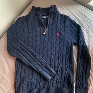 Marinblå stickad tröja Polo Ralph Lauren - Säljer en marinblå stickad tröja från Polo Ralph Lauren med klassiskt kabelstickat mönster och röd logga på bröstet. Tröjan har half zip och hög krage, samt långa ärmar och ribbade muddar. Fick i present så vet inte om den är äkta. Storlek M-L