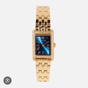 Guldig klocka från Ivy N Rox med blå urtavla - Snygg guldig klocka från Ivy N Rox med rektangulär form, blå urtavla och romerska siffror. Klockan har länkarmband i metall och glittriga stenar runt urtavlan som ger extra bling. Perfekt accessoar för att lyfta din outfit. Aldrig använd. 