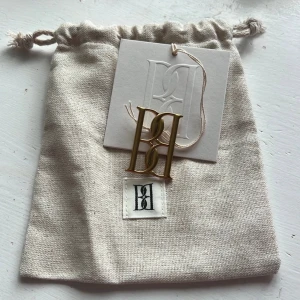 Guld brosch från By Malene Birger - Snygg brosch i guldtonad metall från By Malene Birger i storlek small. Formad som deras ikoniska monogram. Perfekt att fästa på halsduk, kappa eller tröja för en stilren och lyxig touch. Kommer med originalpåse och etikett.