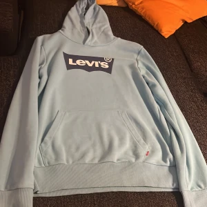 Ljusblå Levi's hoodie med huva - Säljer en ljusblå hoodie från Levi's med stor logga framtill. Tröjan har huva, magficka och ribbade muddar vid ärmslut och nederkant. Perfekt för chill dagar och enkel att matcha med jeans eller joggers.