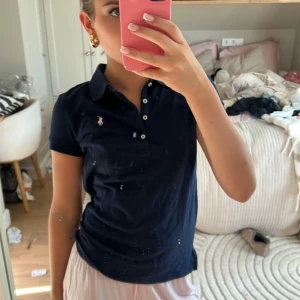 Marinblå pikétröja från Ralph Lauren - Klassisk marinblå pikétröja från Ralph Lauren med vit broderad logga på bröstet. Tröjan har korta ärmar, krage och knappar framtill. Perfekt för en sportig och stilren look.