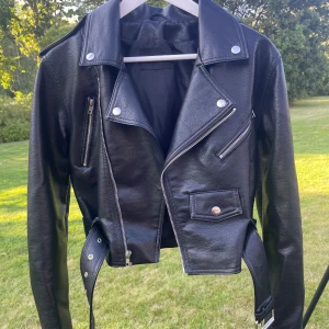 Svart skinnjacka från BikBok, S - Snygg svart skinnjacka från BikBok i bikerstil med silvriga dragkedjor, knappar och bälte i midjan. Jackan har klassisk krage, flera fickor och är croppad för en cool look. Perfekt för dig som vill ha en edgy vibe till din outfit.