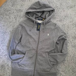 Grå zip-hoodie från Polo Ralph Lauren - Snygg grå hoodie från Polo Ralph Lauren med dragkedja och huva. Tröjan har långa ärmar, snörning vid huvan och två praktiska fickor framtill. Klassisk logga broderad på bröstet. Aldrig använd, endast provad. Äkta såklart (bild 5). Nyskick. Hör av er vid frågor!😀