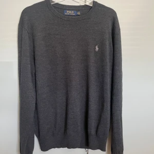 100 % merinoullströja Polo Ralph Lauren - Snygg mörkgrå tröja från Polo Ralph Lauren i mjuk merinoull. Klassisk rund halsringning, diskret broderad logga på bröstet och ribbade muddar. Perfekt för dig som gillar stilren och enkel design med premiumkänsla.