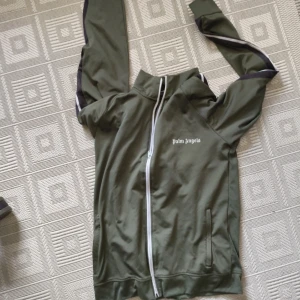 Grön vindjacka från Palm Angels - Snygg grön vindjacka från Palm Angels med hel dragkedja och vita detaljer. Jackan har svarta och vita ränder längs ärmarna och en hög krage. Perfekt för en sportig och trendig look.