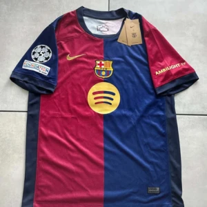 FC Barcelona Lamine Yamal matchtröja - Snygg FC Barcelona matchtröja med Lamine Yamal och nummer 19 på ryggen. Tröjan är röd och blå med guldfärgade detaljer, klubbmärke och sponsorer. Korta ärmar, rund hals och tillverkad i lätt, ventilerande material. Perfekt för dig som älskar Barca och vill sticka ut på läktaren.