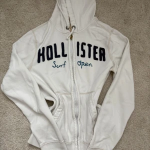 Vit hoodie från Hollister - Säljer en vit hoodie från Hollister med mörkblå broderad logga och texten 'Surf Open' framtill. Tröjan har huva med snörning, dragkedja och två fickor.