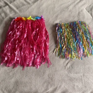 Två färgglada Hawaiikjolar - Säljer två Hawaiikjolar i syntetmaterial, perfekta för temafest eller midsommar. Ena kjolen är lång och knallrosa med blommor i midjan, den andra är kort och har flerfärgade glansiga remsor. Båda har elastisk midja för skön passform.