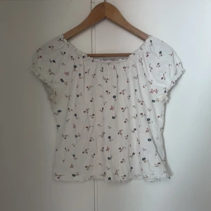 Vit blommig offshoulder blus - Supersöt vit blus med små blommor i rosa och blått. Offshoulder-modell med resår upptill och korta ärmar med volangkant. Det finns gula fläckar på tröjan, finns bild på det. Strl M