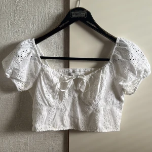 Vit broderad blus från Hollister - Superfin vit blus från Hollister med broderade detaljer och puffärmar. Toppen är croppad, har knyt framtill och är gjord i bomull med spets och broderade hålmönster. Perfekt för sommarens alla tillfällen och ger en romantisk vibe.