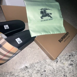 Burberry sandaler med klassiskt rutmönster - Snygga Burberry sandaler med den ikoniska beigea och svarta rutan på remmen. Svart sula med präglad Burberry-logga. Tillverkade i Italien och levereras med originalkartong och dustbag. Perfekta för sommaren och ger en lyxig touch till din outfit.