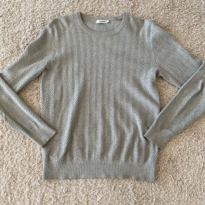 Grå stickad tröja från J.Lindeberg - Snygg grå stickad tröja från J.Lindeberg med ett stilrent fiskbensmönster över hela plagget. Rund halsringning och långa ärmar. Perfekt för dig som gillar klassisk men modern stil. Materialet är mjukt och skönt mot huden.