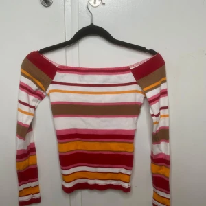 Randig offshoulder tröja från BikBok - Säljer en ribbad offshoulder tröja från BikBok i storlek XS. Tröjan har lång ärm och färgglada ränder i vitt, rött, rosa, gult och beige. Den är slim fit och har en mjuk, stretchig känsla som sitter snyggt på kroppen. Perfekt för dig som gillar färg.