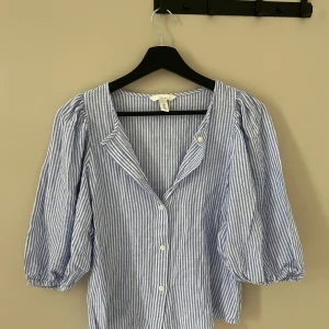 Blårandig linneblus från H&M - Snygg blårandig blus från H&M i linneblandning. Skjortan har puffiga trekvartsärmar med resår vid ärmslut, klassisk krage och knappar framtill. Perfekt för dig som gillar en chill och fräsch stil med lite volym i ärmarna.