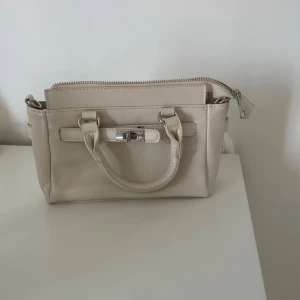 Beige handväska med axelrem - Snygg beige handväska i skinnimitation med både handtag och justerbar axelrem. Väskan har silverfärgade detaljer. Helt oanvänd från stradivarius💞