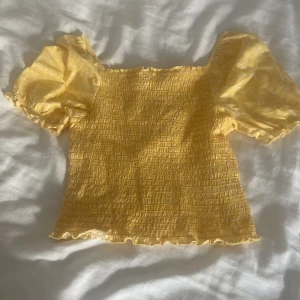 Gul smockad blus från KappAhl - Supersöt gul blus från KappAhl med smockad kropp och puffiga korta ärmar. Ärmarna har små vita blommor och volangkant, medan resten av toppen är ribbad och har en lätt croppad passform. Perfekt för soliga dagar! 