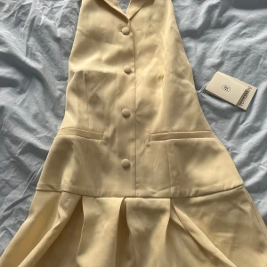 Champagnefärgad kortklänning från Peppermint - Supersnygg kortklänning i champagnefärg från Peppermint med krage och knappar framtill. Klänningen har en omlottliknande halterneck, två fickor och ett plisserat kjolparti. Perfekt för dig som gillar stilrena och trendiga detaljer.