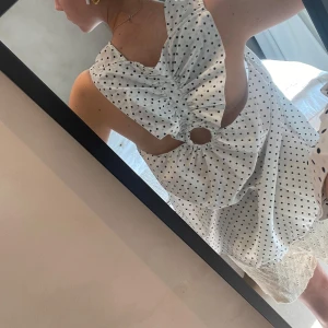 Vit prickig ärmlös blus med öppen rygg - Superfin vit blus med svarta prickar och en unik öppen rygg med snygg rynkdetalj och rund utskärning. Ärmlös och luftig modell som passar perfekt till varma dagar. Ger en cool och trendig vibe till din outfit.