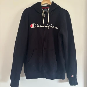 mörkblå Champion hoodie med dragkedja - mörkblå hoodie från Champion i storlek medium med vit snörning och broderad logga på bröstet. Tröjan har huva, fickor framtill och dragkedja hela vägen. Mjuk insida och klassisk passform, perfekt för chill eller streetwear.