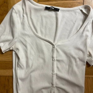 Vit ribbad topp med knappar Bik Bok - Superfin vit ribbad topp från Bik Bok med korta ärmar och v-ringning. Toppen har knappar hela vägen framtill och är gjord i mjuk bomull. Perfekt för dig som gillar en enkel men ändå snygg stil. OBS! Lite nopprig i materialet