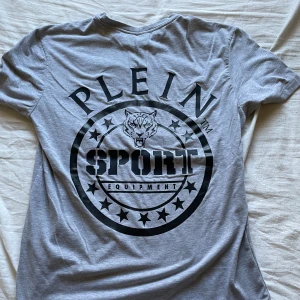 Grå Plein Sport t-shirt med tryck - Snygg grå t-shirt från Plein Sport med stort svart tryck på ryggen och diskret logga framtill. Klassisk passform och rund hals. Perfekt för dig som gillar sportiga och stilrena plagg med attityd. Tillverkad i mjuk bomull för skön känsla hela dagen.