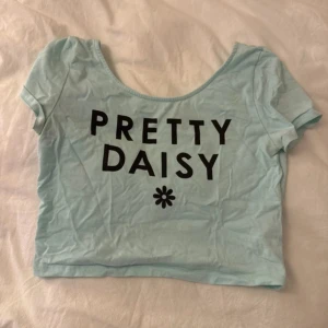 Ljusblå croppad topp Pretty Daisy - Superfin ljusblå croppad topp med svart text 'PRETTY DAISY' och en liten blomma framtill. T-shirten har korta ärmar och rund halsringning. Perfekt för dig som gillar gulliga och trendiga prints.