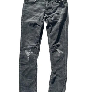 Svarta Nudie Jeans med slitningar - Säljer ett par snygga nudie jeans med slitningar från fabrik. Jeansen stängs med snappar. Byxorna är i storlek W29, L32. Byxorna är gjorda av hårt tyg. Tyvärr är det en liten skada (se bild nr5). Skadan är inte slitage. Byxorna är av väldigt bra kvalitet och känns som ett äkta par jeans! Ta gärna kontakt vid eventuella frågor eller bilder😊. 
