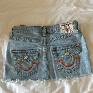 True Religion ljusblå jeanskjol - Säljer en ljusblå kort jeanskjol från True Religion, modell Sadie Mini Skirt. Kjolen har fransad nederkant, klassiska fickor med broderade detaljer i rött och gult, samt silverfärgade knappar. Perfekt för dig som gillar street och denim vibes.