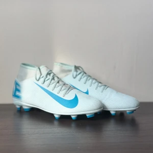Nike Mercurial fotbollsskor vit/blå - Nike Mercurial fotbollsskor i storlek 45.5, med en clean vit bas och ljusblå detaljer. Skorna har snörning, platt sula med dobbar och en snygg Nike-logga på sidan och hälen. Ovandelen är i syntetmaterial för lätt känsla och bra grepp på planen.
