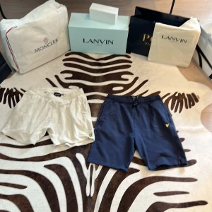 Lyle & Scott shorts i frotté och bomull - Två par snygga shorts från Lyle & Scott, ett par i ljus frotté och ett par i mörkblå bomull. Båda har resår i midjan och den klassiska gula fågel-loggan broderad framtill. Perfekta för sommaren och chill dagar.