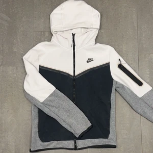Nike hoodiejacka med dragkedja - Nike tech fleece i vitt, svart och grått med hel dragkedja framtill och huva. Koftan har en snygg färgblock-design, en praktisk ficka med dragkedja på ärmen och Nike-logga på bröstet. Perfekt för chill dagar eller när du vill ha en clean look