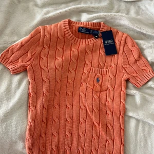 Stickad orange tröja Polo Ralph Lauren - Säljer en snygg stickad tröja från Polo Ralph Lauren i en fräsch orange färg. Tröjan har korta ärmar, rund hals och ett klassiskt kabelstickat mönster. På bröstet finns en liten ficka med den ikoniska blå Polo-loggan. Perfekt för vår och sommar! Pris lappen är från Qatar så det står i Qatar pengar. Den kostar egentligen 1500. 