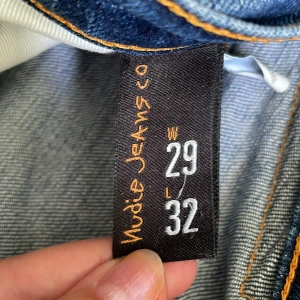  Nudie Jeans, lean Dean, W29 L32 - Säljer ett par klassiska blå jeans(lean Dean)från Nudie Jeans i storlek W29 L32. Jeansen har snygga slitningar, orange kontrastsömmar och den ikoniska brodyren på bakfickan. Modellen har rak passform och är tillverkad i mjukt denimtyg av bomull.