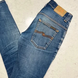 Blå jeans från Nudie Jeans, 30/32 - Säljer ett par jeans från Nudie. Slim fit med ett hål/slitning under höger knä. Perfekt till ett par snygga sneakers. Skulle säga att storleken är lite mindre än vad som anges. Hör av dig vid fler frågor!