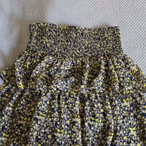 Volangkjol  - NY utan prislapp! Supersöt kortkjol från Vero Moda med blommigt mönster i gult, rosa och lila på mörkblå botten. Kjolen har smockad midja och volanger som ger en flowy känsla. Perfekt volang sommar kjol! Går att ha low waist och highwaist och går att vika ner för att få den ännu mer low waist! Passar S-L då den är stretchig! Skriv om du har några frågor eller om du är intresserad! Pris kan diskuteras! 