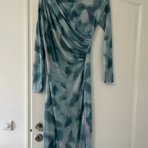 Blå långklänning med drapering - Säljer en långklänning i blå och grå toner med abstrakt mönster. Klänningen har lång ärm, omlott-drapering över axel och midja samt en snygg rynkning på sidan. Materialet känns mjukt och följsamt, perfekt för dig som gillar en cool och unik look.